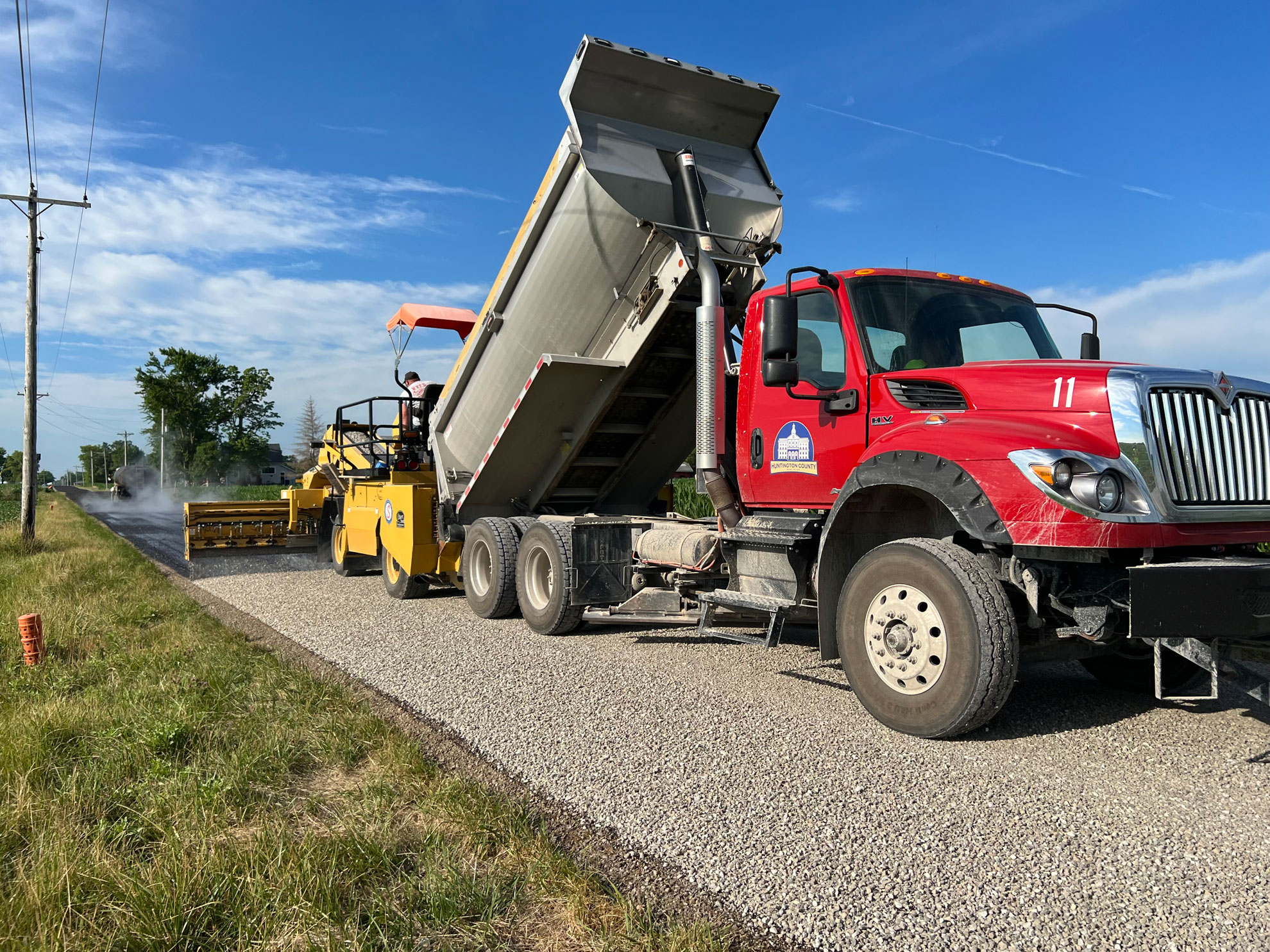 A dump truck and paver work in tandem to chip and seal a road
