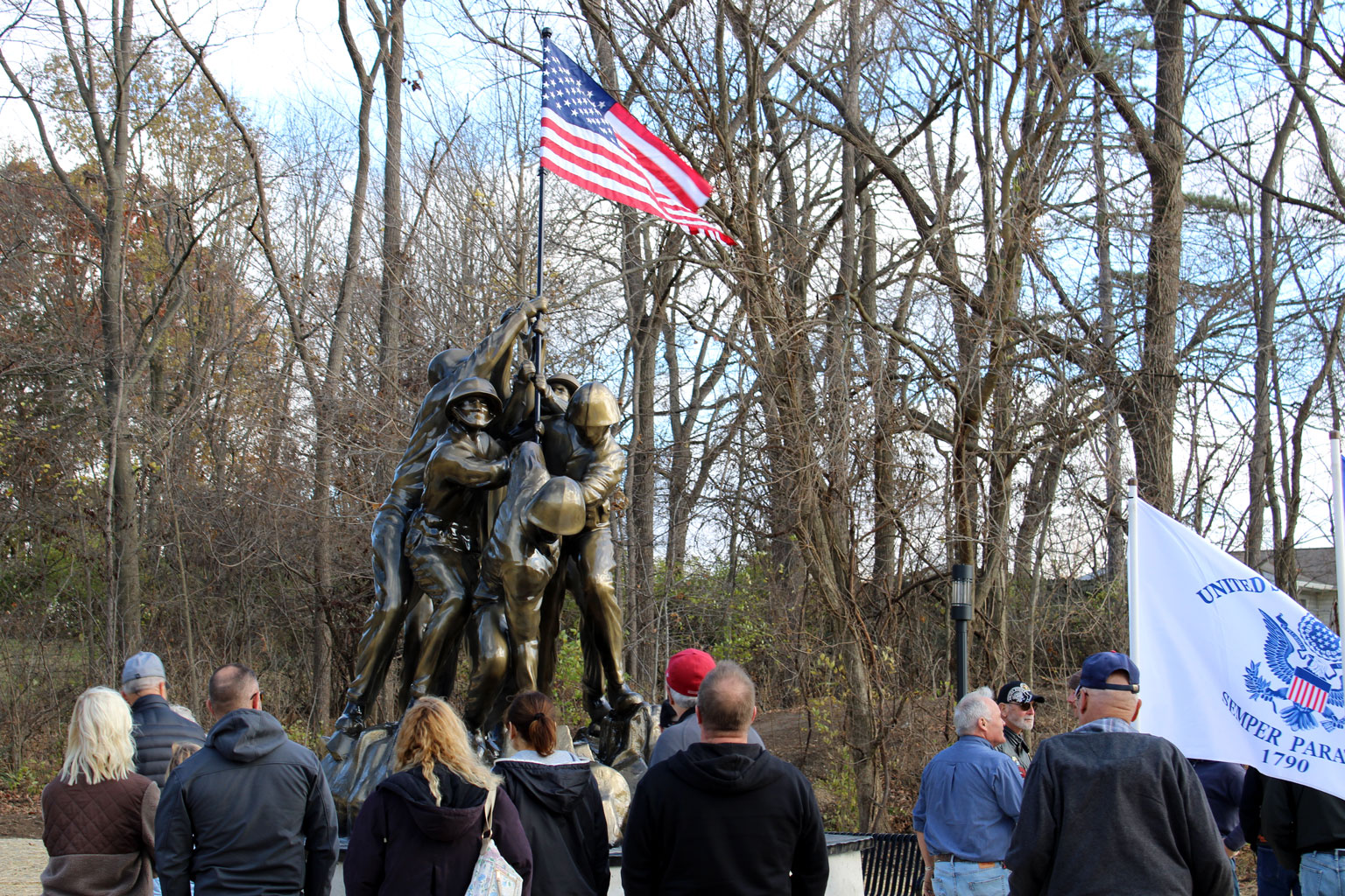 The Iwo Jima Memorial in Memorial Park, Huntington, Indiana, unveiled in November 2024. The bronze figures raise the American flag against a clear sky, surrounded by visitors at the dedication ceremony.